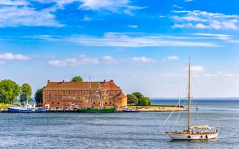 Schloss Sonderburg, ein großes Backsteingebäude mit orangefarbenem Ziegeldach, am Wasser. Ein Segelboot fährt im Vordergrund auf blauem Wasser.