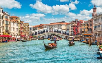 Die berühmte Rialtobrücke überspannt den Canal Grande in Venedig mit Gondeln und Booten im Wasser. Bunte historische Gebäude säumen beide Seiten unter blauem Himmel.