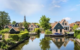 Ruhiger Kanal in Hindeloopen mit traditionellen niederländischen Häusern, roten Ziegeldächern und Booten am Ufer. Ein Kirchturm erhebt sich im Hintergrund.
