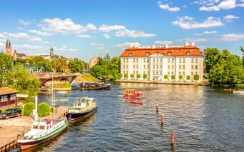 Schloss Köpenick mit orangefarbenem Dach neben einem Fluss in Berlin. Boote am Ufer vertäut, Brücke und Kirchtürme im Hintergrund sichtbar.