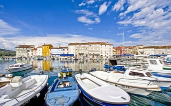 Hafen auf der Insel Cres mit bunten Booten im ruhigen Wasser. Mediterrane Gebäude säumen die Uferpromenade unter blauem Himmel mit weißen Wolken.