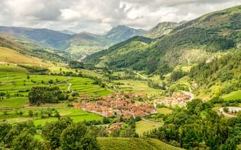 Luftaufnahme eines Dorfes mit Terrakottadächern in einem grünen Tal, umgeben von bewaldeten Bergen unter bewölktem Himmel in Spanien.