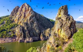 Dramatische Felsformationen am Castillo de Monfragüe, die aus einem Stausee aufragen. Vögel kreisen über den Klippen unter blauem Himmel.