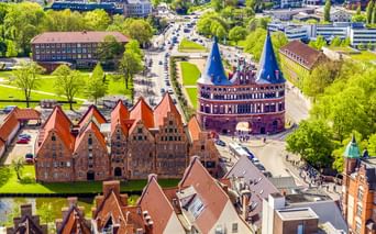 Luftaufnahme von Lübeck mit dem ikonischen Holstentor mit Zwillingstürmen und roten Backsteingebäuden, umgeben von grünen Parks und Stadtgebieten.