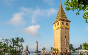 Historischer Mangturm mit buntem Ziegeldach am Lindauer Hafeneingang am Bodensee. Leuchtturm im Hintergrund, Palmen im Vordergrund.
