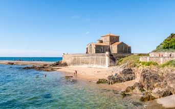Kirche Prieuré Saint-Nicolas an felsiger Küste in Sables-d'Olonne mit Sandstrand, klarem blauen Wasser und Strandbesuchern unter sonnigem Himmel.