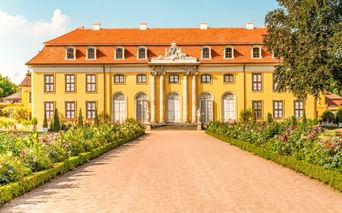 Gelbes Barockschloss mit orangefarbenem Dach und weißen Säulen in Mosigkau bei Dessau. Ein Kiesweg führt durch einen formalen Garten zum Eingang.