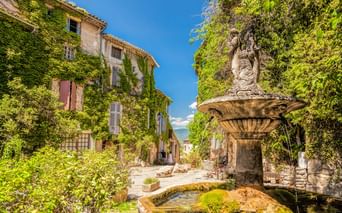 Steinbrunnen mit Statue auf dem Dorfplatz von Saignon, umgeben von efeubedeckten Gebäuden und üppigem Grün unter blauem Himmel.