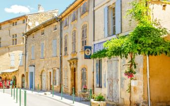 Sonnige Straße in Aix-en-Provence mit traditionellen Steingebäuden, pastellfarbenen Fensterläden und grünen Bäumen. Grüne Poller säumen den Fußgängerbereich.