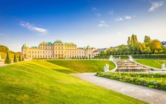 Schloss Belvedere in Wien mit gepflegten Barockgärten, weißen Statuen, Brunnen und grünen Rasenflächen unter blauem Himmel.