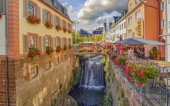 Wasserfall in Saarburg zwischen bunten historischen Gebäuden mit Blumenkästen. Straßencafés mit Sonnenschirmen säumen die Straße über dem Wasserfall.