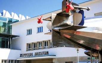 Weißes modernistisches Gebäude mit Zeppelin Museum Eingangsschild und großer Metall-Propeller-Skulptur mit roten Akzenten über dem Eingang.
