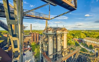 Erhöhte Ansicht historischer Hochöfen und Industriestrukturen im Landschaftspark Duisburg-Nord, umgeben von grüner Vegetation unter blauem Himmel.