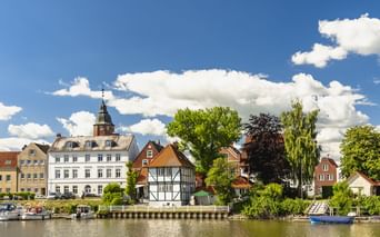 Uferblick auf Glückstadt mit historischen Gebäuden, darunter ein weißer Kirchturm, Fachwerkhaus und Boote am Hafen unter blauem Himmel.