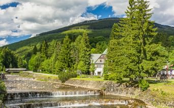 Kaskadenartige Stromschnellen der Labe in Spindlermühle mit Steinterrassen, umgeben von grünen Bäumen und einem weißen Haus, Berge im Hintergrund.