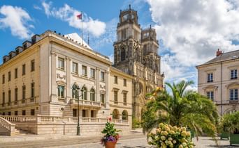 Gotische Zwillingstürme der Kathedrale Sainte-Croix in Orléans hinter einem cremefarbenen Gebäude mit Palmen und Blumen auf dem Platz.