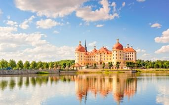 Schloss Moritzburg mit gelber Fassade und roten Kuppeln spiegelt sich im umgebenden See unter blauem Himmel mit weißen Wolken.