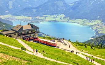 Rote Zahnradbahnen an der Schafberg-Bergstation mit Blick auf türkisfarbenen See und grüne Täler mit Alpengipfeln im Hintergrund.