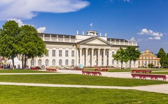 Das neoklassizistische Fridericianum in Kassel mit Säulenfassade, gesehen über eine grüne Rasenfläche mit roten Bänken unter blauem Himmel.
