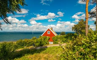 Traditionelles rotes Schwedenhaus mit weißen Verzierungen auf einem Küstenhang, umgeben von grüner Vegetation und Bäumen, mit Blick auf das blaue Meer unter bewölktem Himmel.