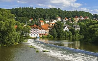 Blick auf Melsungen an der Fulda mit einem Wehr im Vordergrund. Fachwerkhäuser und rote Dächer schmiegen sich zwischen grüne Bäume unter blauem Himmel.