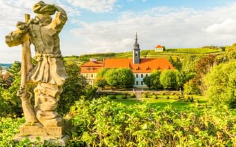 Steinstatue im Vordergrund mit Blick auf Gärten und historische Gebäude mit orangen Dächern in Diesbar-Seußlitz, dahinter weinbergbedeckte Hügel.