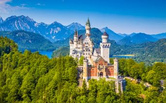 Schloss Neuschwanstein umgeben von grünen Wäldern mit bayerischen Alpen und blauen Seen im Hintergrund unter klarem Himmel.