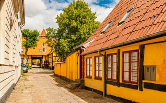 Kopfsteinpflastergasse in Nakskov mit gelben Fachwerkhäusern mit roten Fenstern und Türen, roten Ziegeldächern und einem Torturm im Hintergrund.