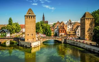 Zwei mittelalterliche Steintürme des Barrage Vauban im Wasser, verbunden durch eine Brücke in Straßburg. Historische Gebäude und Kirchturm sichtbar.