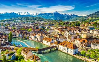 Panoramablick auf Luzerns Altstadt mit bunten Gebäuden entlang der türkisfarbenen Reuss, Holzbrücke und schneebedeckten Alpen im Hintergrund.