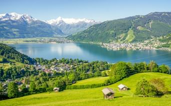 Panoramablick auf den Zeller See umgeben von grünen Hügeln und schneebedeckten Alpengipfeln. Traditionelle Holzhütten auf Wiesen im Vordergrund.