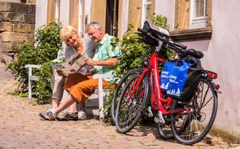 Zwei Radfahrer sitzen auf einer weißen Bank und lesen eine Karte in Bad Wimpfen. Zwei rote Tourenräder mit blauen Taschen stehen daneben.