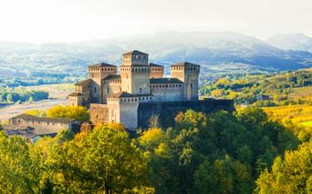 Burg Torrechiara auf einem bewaldeten Hügel, umgeben von grünen Bäumen mit sanften Hügeln und Bergen im Hintergrund bei weichem Licht.
