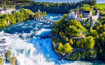 Luftaufnahme des Rheinfalls bei Schaffhausen mit kraftvollem Wasserfall, historischem Schloss, Steinbrücke und grünen Wäldern unter blauem Himmel.