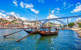 Traditionelle Rabelo-Boote mit Weinfässern auf dem Douro in Porto, mit der Dom-Luís-I-Brücke und bunten Häusern am Hang im Hintergrund.