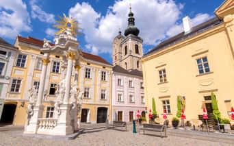Barocker Platz in Krems-Stein mit verziertem weißen Monument, pastellfarbenen Gebäuden und Kirchturm unter blauem Himmel mit weißen Wolken.