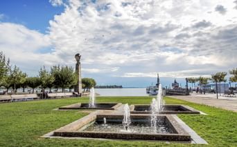 Stufenbrunnen mit Wasserfontänen auf grüner Wiese am Hafen in Konstanz. Bodensee und historisches Hafengebäude im Hintergrund unter bewölktem Himmel.