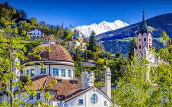 Blick auf Meran mit Kuppel des Kurhauses, Kirchturm mit Uhr und schneebedeckten Bergen. Frühlingslaub rahmt die historischen Gebäude unter blauem Himmel.