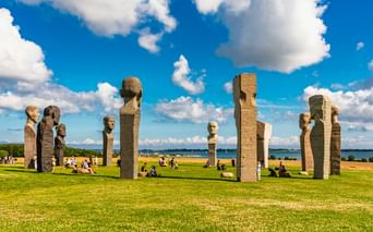 Dodekalitten-Skulptur bei Kragenæs mit hohen Steinsäulen mit geschnitzten Gesichtern auf grüner Wiese, Besucher entspannen sich, blauer Himmel mit Wolken.