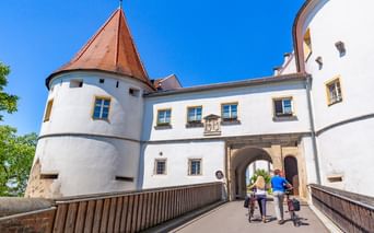 Zwei Radfahrer schieben ihre Räder über eine Brücke zum Eingang von Schloss Wörth. Weiße Schlossmauern mit rotem Turmdach unter blauem Himmel.