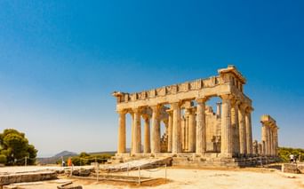 Antiker Aphaia-Tempel auf Ägina mit dorischen Säulen unter klarem blauen Himmel. Besucher erkunden die archäologische Stätte mit Bergen im Hintergrund.