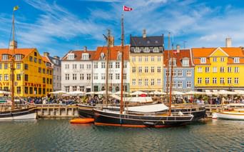 Historische Holzsegelschiffe am Nyhavn-Hafen in Kopenhagen mit bunten gelben, weißen und orangen Stadthäusern entlang der Uferpromenade.