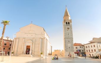Sonniger Hafenplatz in Umag, Kroatien, mit weißer Kirche und steinernem Glockenturm. Palme links, Fußgänger auf dem Platz unter blauem Himmel.