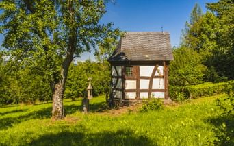 Fachwerkkapelle mit weißen Wänden und braunen Balken im Freilichtmuseum Roscheider Hof in Konz, umgeben von grüner Wiese und Bäumen unter blauem Himmel.