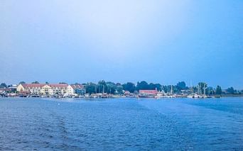 Blick über ruhiges blaues Wasser auf einen Hafen der Insel Poel mit Segelbooten, Wohngebäuden mit roten Dächern und Bäumen unter klarem Himmel.
