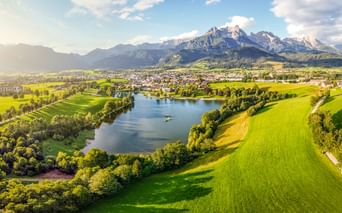 Luftaufnahme von Saalfelden mit einem See, umgeben von grünen Wiesen und Wäldern. Berge erheben sich im Hintergrund unter blauem Himmel mit Wolken.