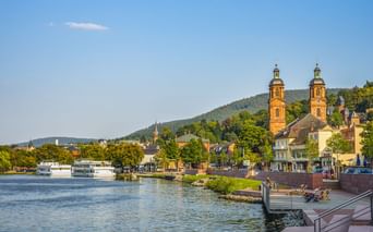 Blick auf Miltenberg mit den Zwillingstürmen der St. Jakobus Kirche am Main. Uferpromenade mit Booten, Hügel im Hintergrund.