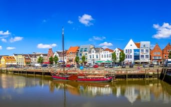 Historisches hölzernes Segelboot am Husumer Hafen mit bunten traditionellen Gebäuden entlang der Uferpromenade unter blauem Himmel.
