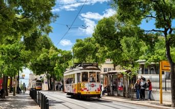 Gelbe historische Straßenbahn 28 auf baumgesäumter Straße in Lissabon mit wartenden Fahrgästen unter blauem Himmel mit weißen Wolken.