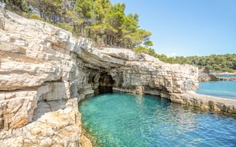 Natürliche Kalksteinhöhle mit türkisfarbenem Wasser in einer Bucht bei Pula. Weiße geschichtete Felsformationen mit grünen Bäumen unter blauem Himmel.